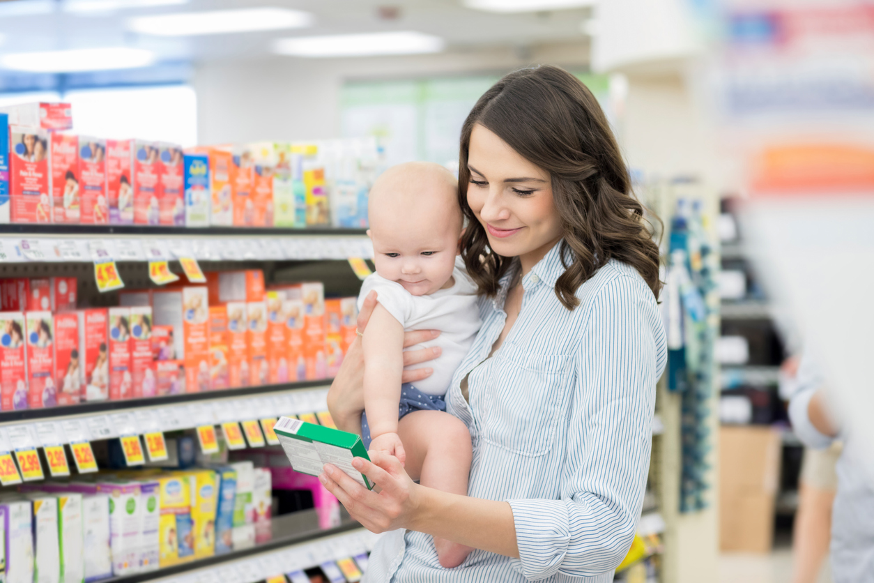 A woman reads the label on a medication while shopping in the pharmacy store, while holding her baby. A woman reads the label on a medication while shopping in the pharmacy store, while holding her baby.